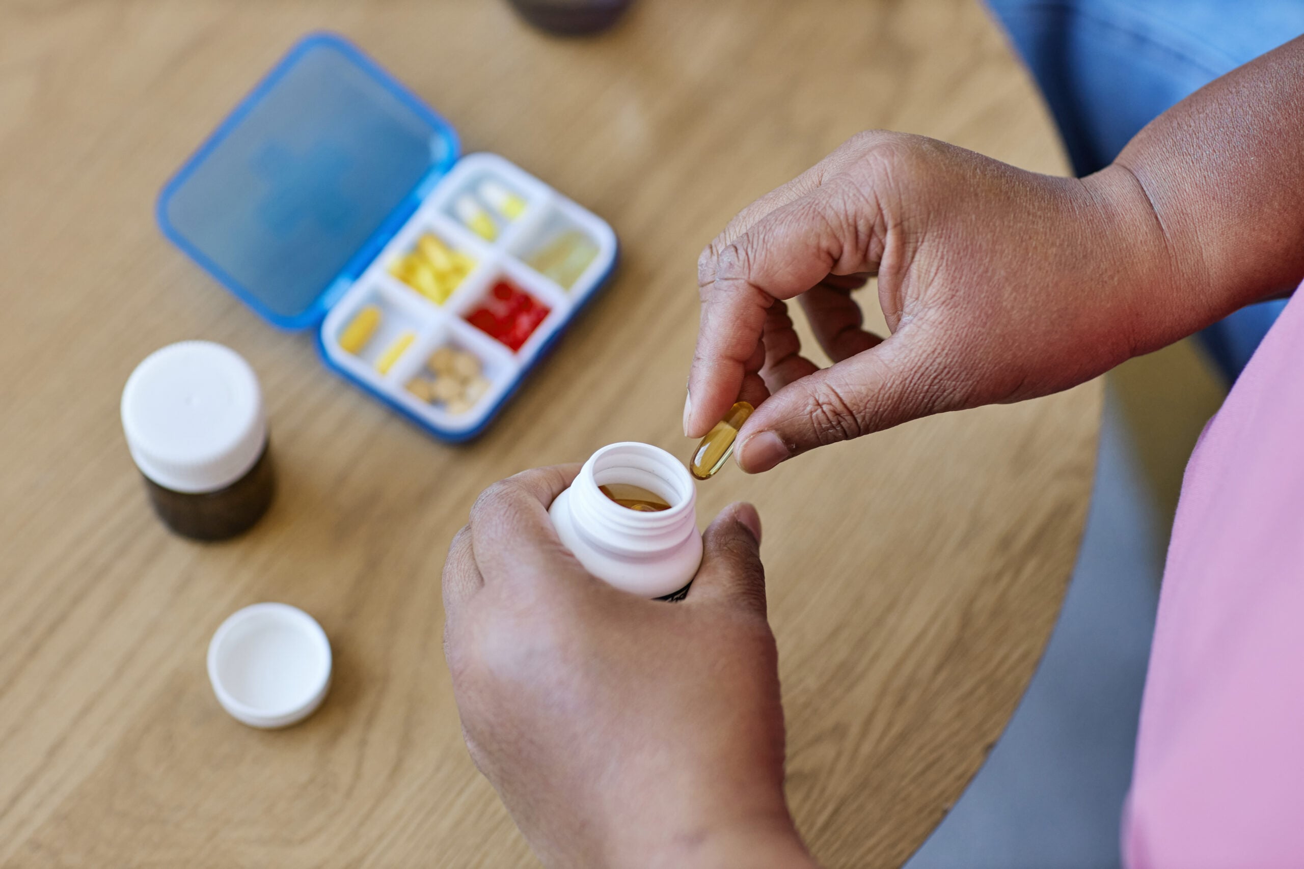 Above view of hands of aged African American woman taking capsule out of white plastic pill-bottle while sitting by table with medicaments