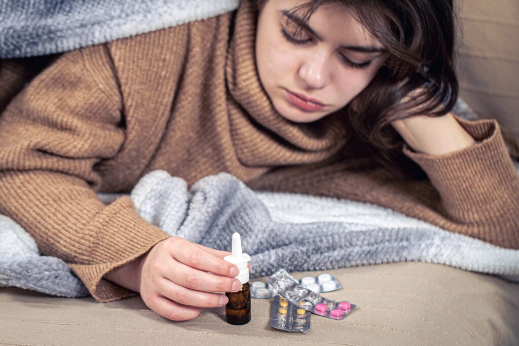Sick young woman with medications lies in bed under a blanket, colds and home treatment.