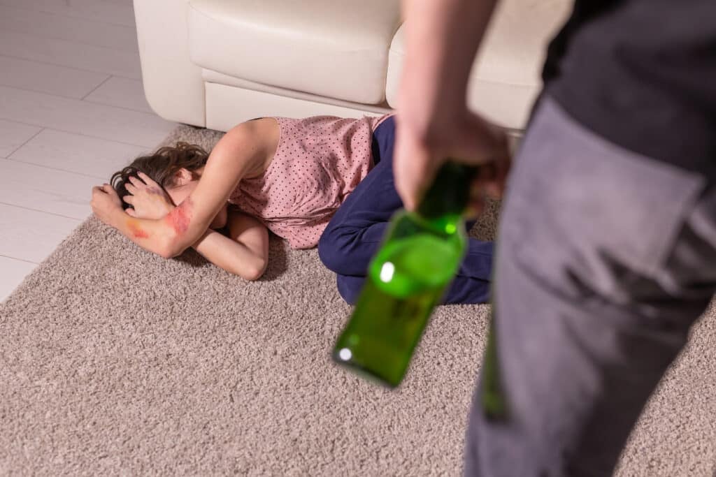 Alcoholism, abuse and domestic violence - Woman lying on the floor. She is afraid of man with bottle
