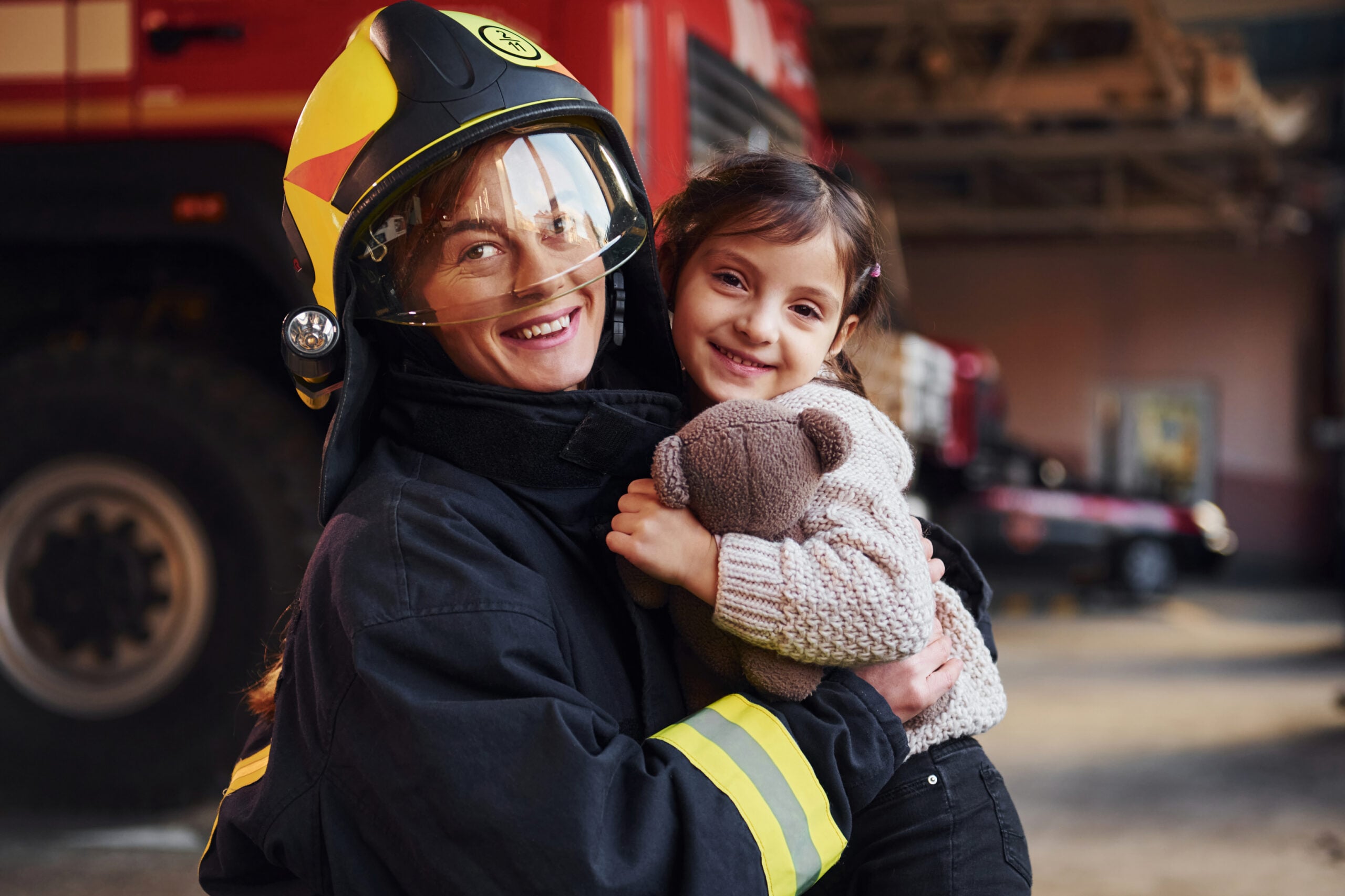 Happy little girl is with female firefighter in protective uniform