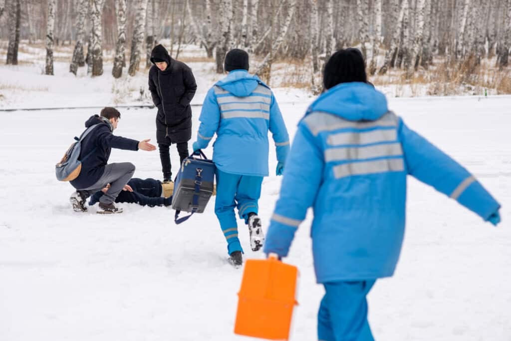 Hypothermia - Rear view of young paramedics in blue uniform hurrying to sick person lying in snow while two guys bending over him