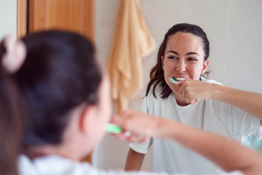 causes of mouth ulcers - miling young woman brushing teeth in bathroom. Happy girl looking in mirror while using ecological toothbrush with whitening toothpaste.