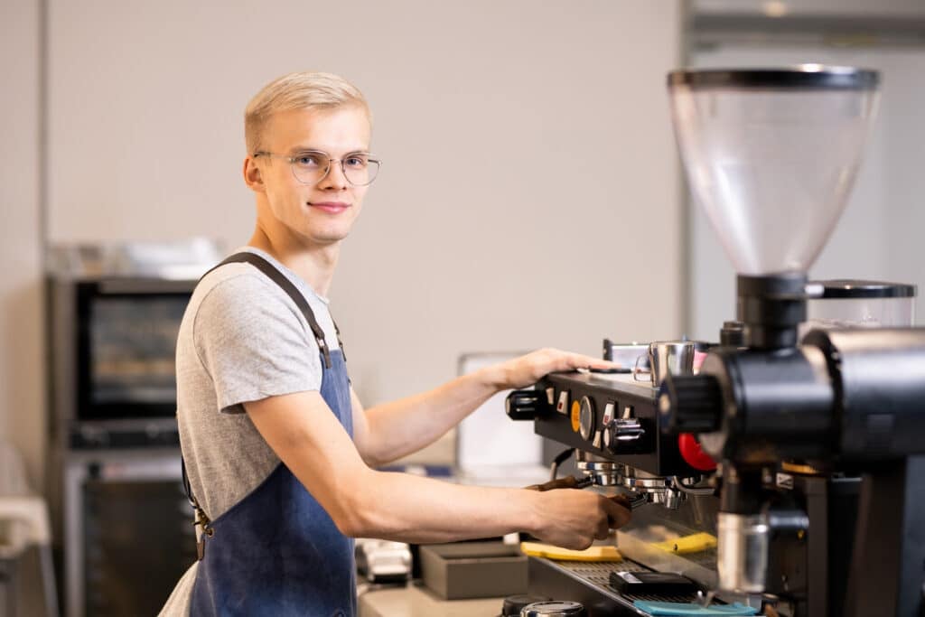 World Youth Skills Day - Young successful barista in uniform using coffee machine while standing by workplace and making fresh coffee for guests