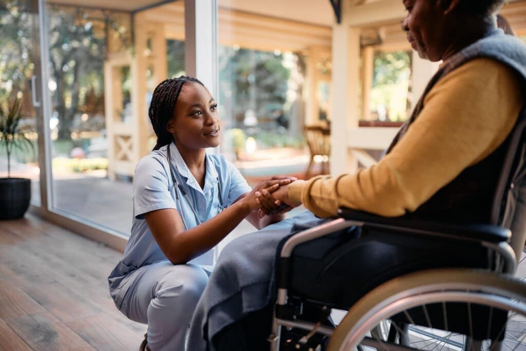 Nurse - Young African American nurse holding hands with senior man in wheelchair while communicating with him at residential care home. Copy space.