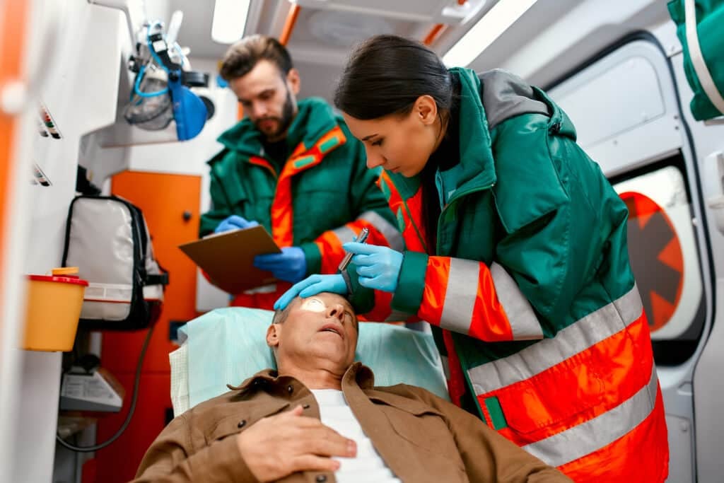 Two paramedics are examining the pupils of a senior lying on a gurney in a modern ambulance and looking at the patient's records.