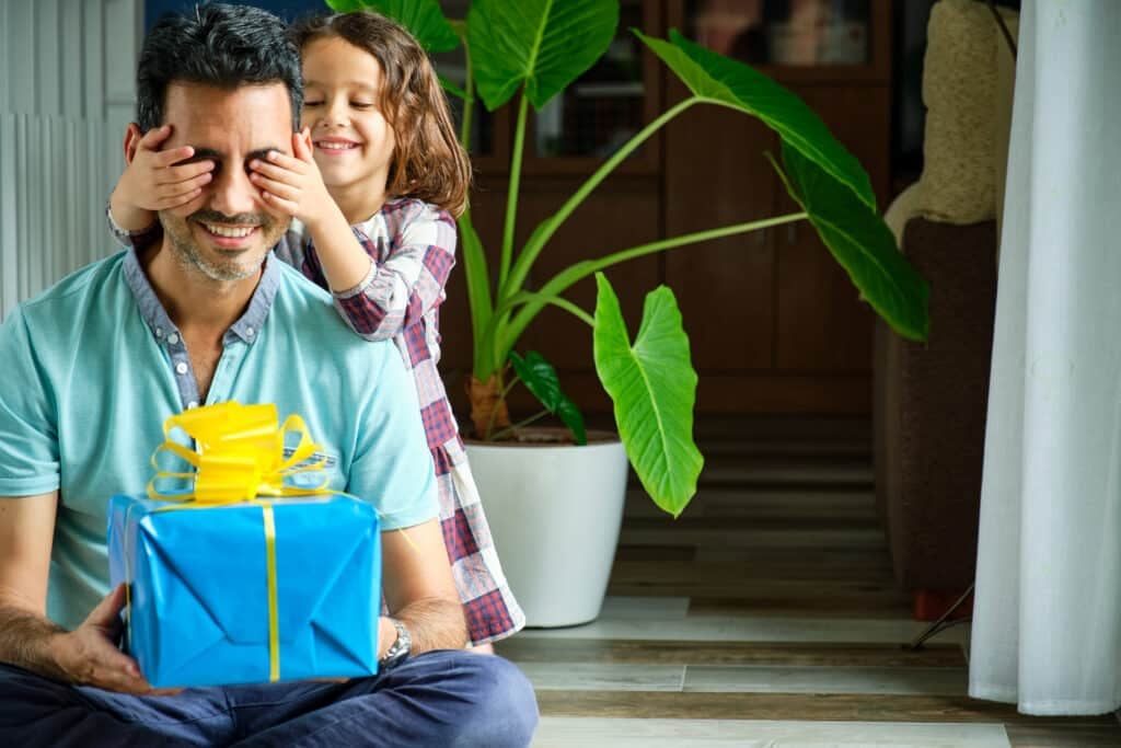little girl covering her eyes to her father who is sitting on the floor with a gift on her legs that she has not seen yet