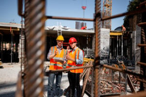 National Safe Work Month 2026 - Architect and structural engineer in orange work vests and helmets discuss a building project on the open air building site with a lot of steel frames